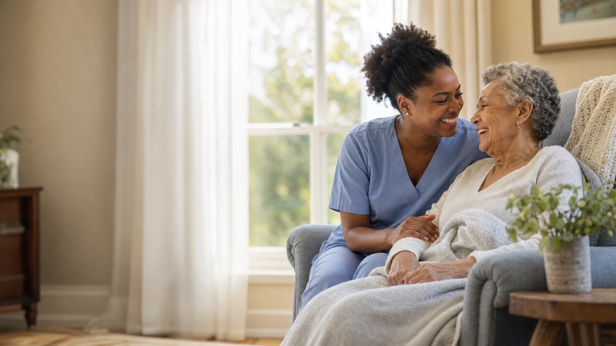 A caregiver in blue scrubs sitting beside an older woman in an armchair by a sunlit window, both laughing